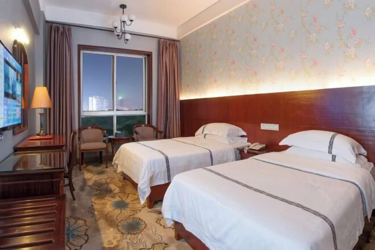 Habitació d'hotel amb dos llits, una taula de nit, una cadira, televisió i finestra amb cortines.