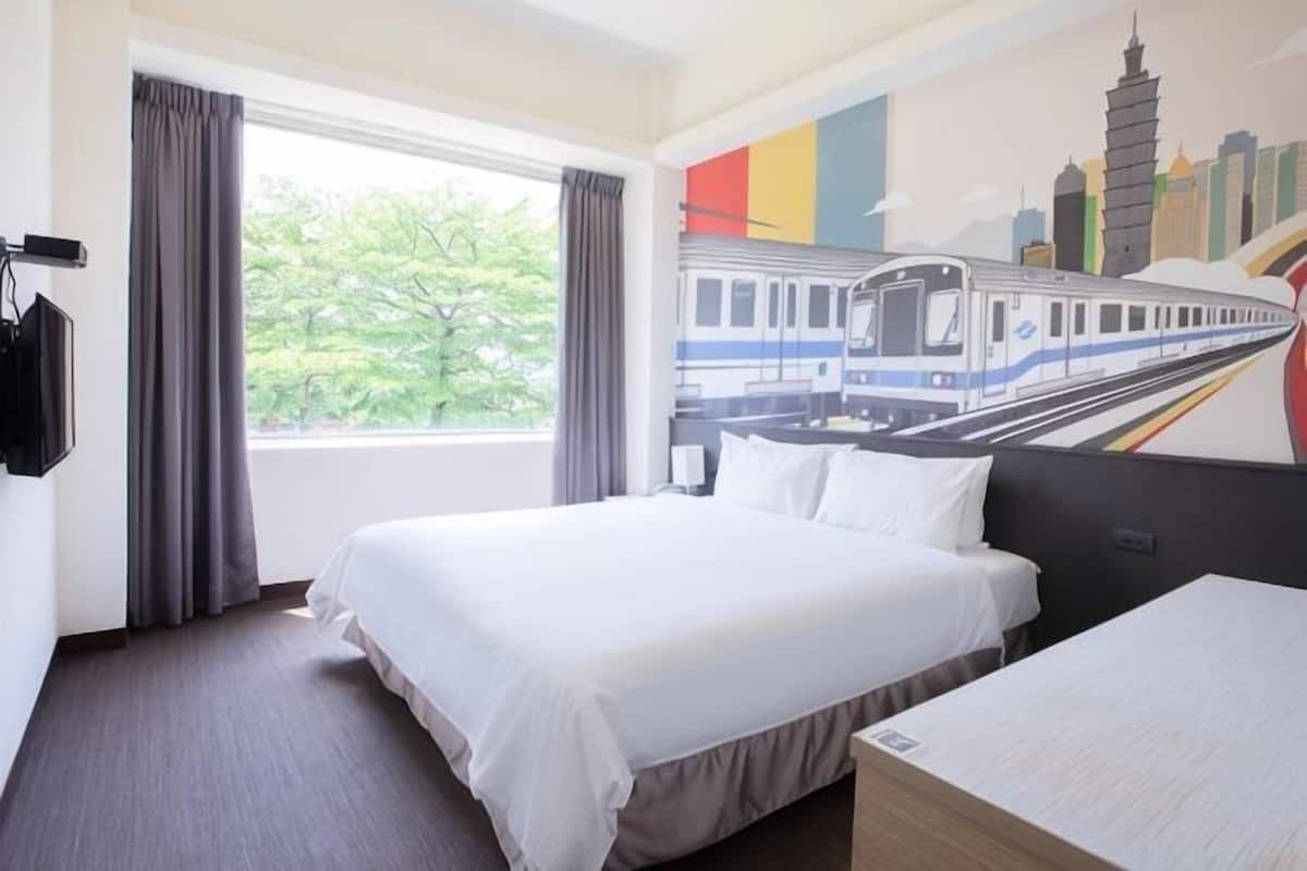 A hotel room with a bed, a desk, a window with curtains, and a cityscape mural on the wall.