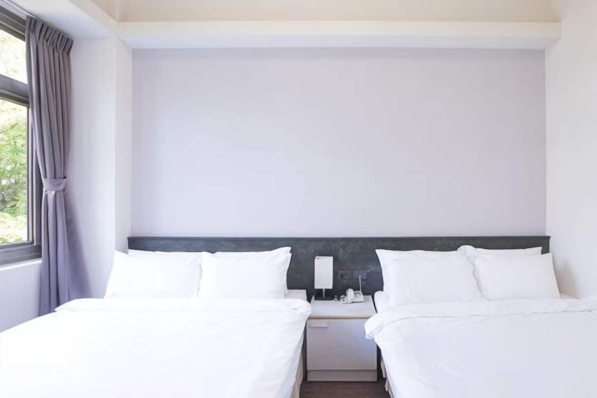 A hotel room with two beds, a nightstand, a lamp, and a window with curtains.