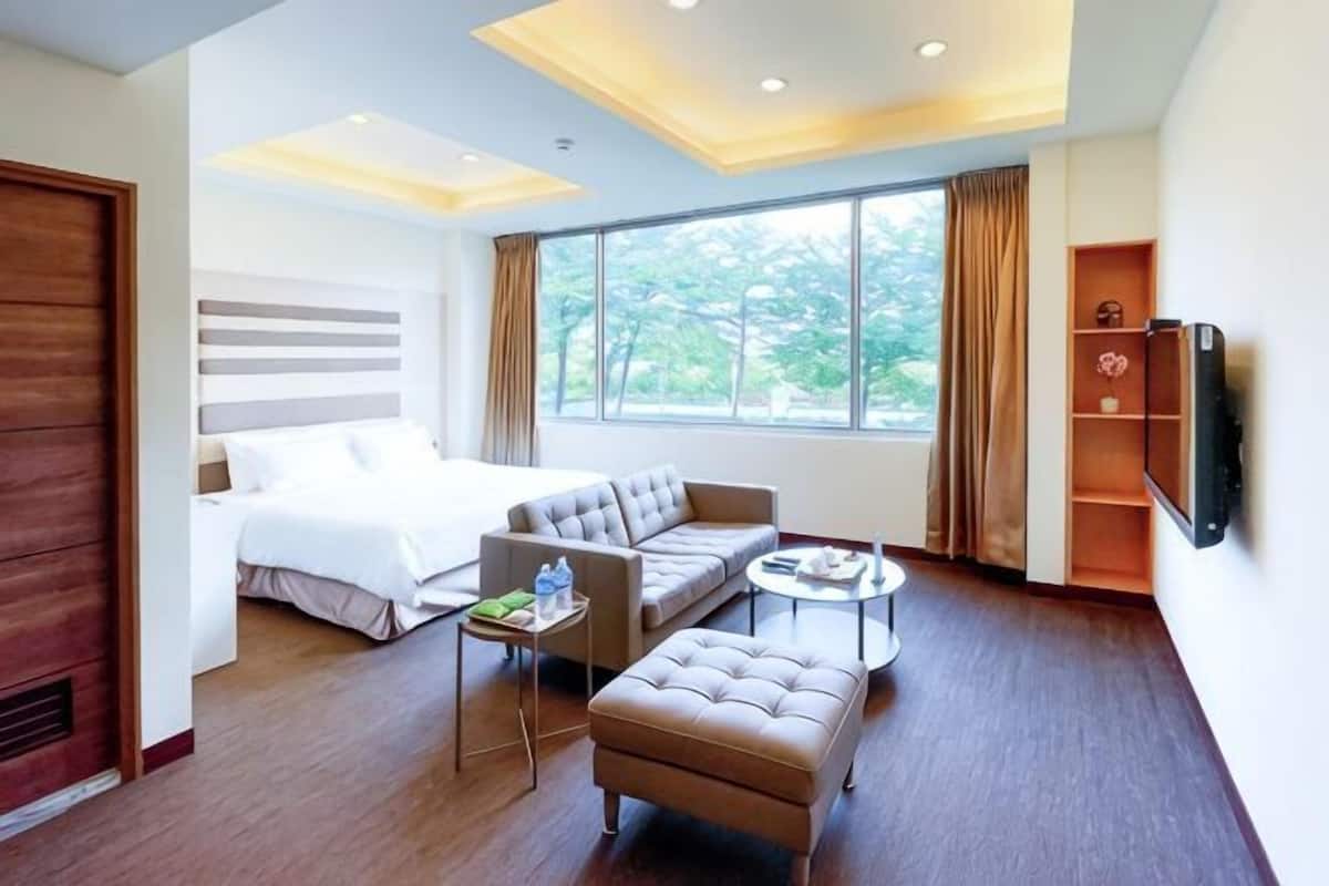 A hotel room with a bed, sofa, ottoman, TV, and a window with curtains.
