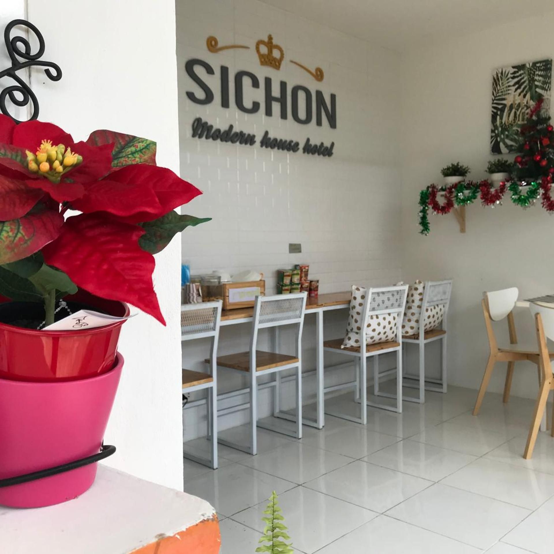 A hotel room with a dining area featuring white chairs and a table. A large potted plant with red flowers is visible.