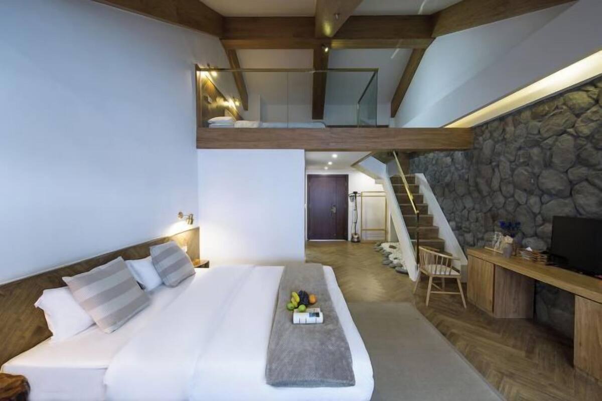 A modern hotel room with a loft bed, a stone wall, a wooden desk, and a staircase.