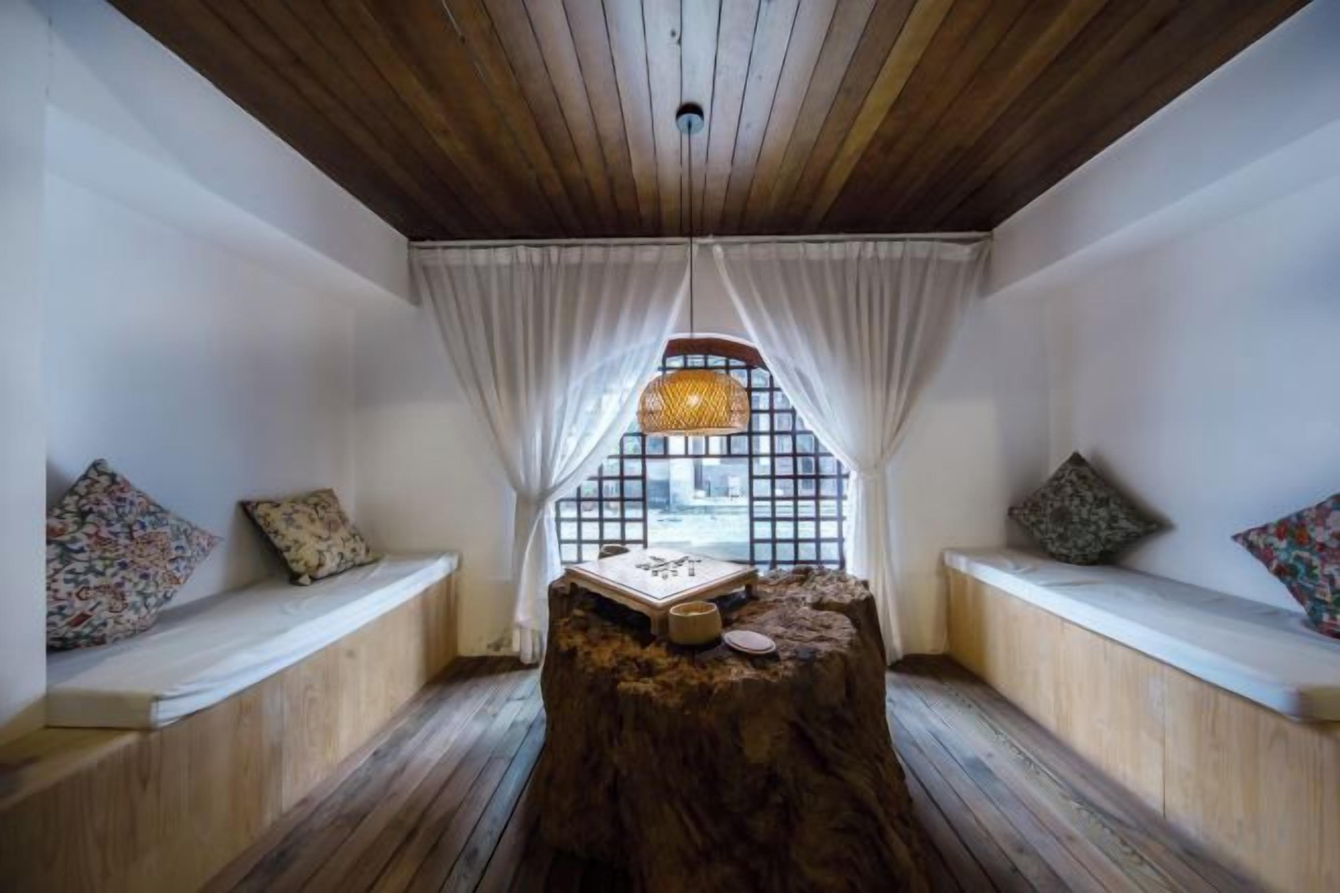 Una habitación con techo de madera, una ventana con cortinas blancas, un banco de madera con cojines y una mesita con una cesta tejida.