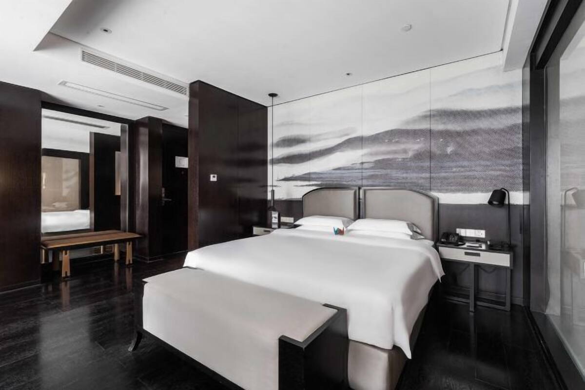 Une chambre d’hôtel moderne avec un grand lit, une table de chevet, un banc et un mur orné d’une fresque représentant un paysage de montagne.