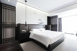 A modern hotel room with a large bed, a headboard with a ship painting, a dark wooden nightstand, a black lamp, and a view of the outdoors through a window with curtains.