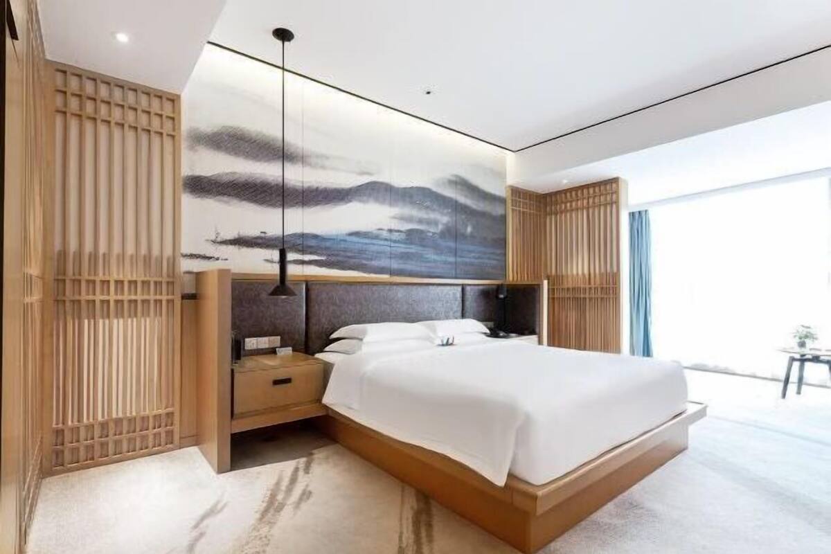 Une chambre moderne avec un grand lit, un lambris en bois, un tableau au mur et une vue sur l’extérieur.