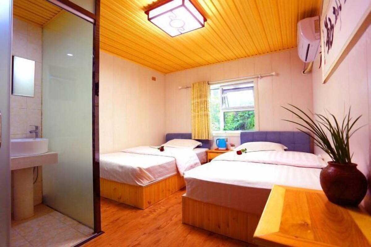 Una habitación con tres camas de madera, una ventana con cortinas, una mesita de noche, una planta y un aire acondicionado instalado en la pared.