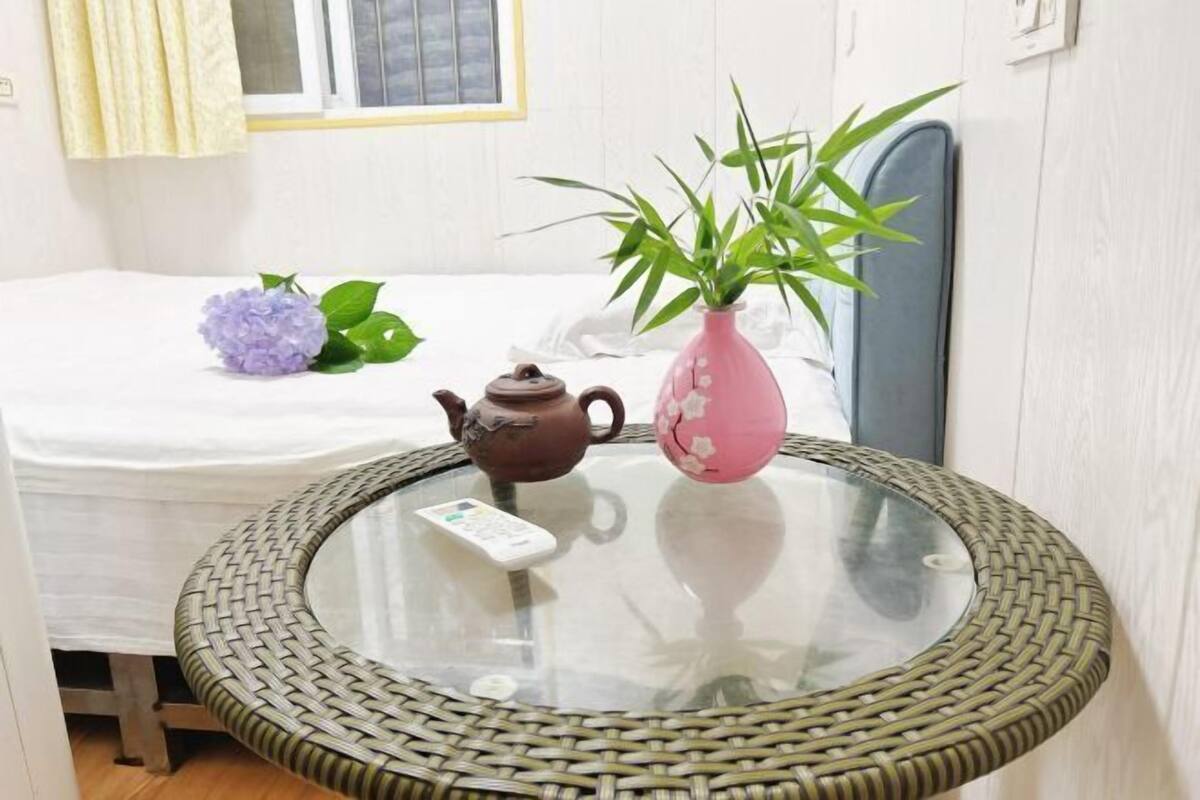 Una mesa redonda de cristal con un borde trenzado, un florero rosa con follaje verde y una tetera marrón.