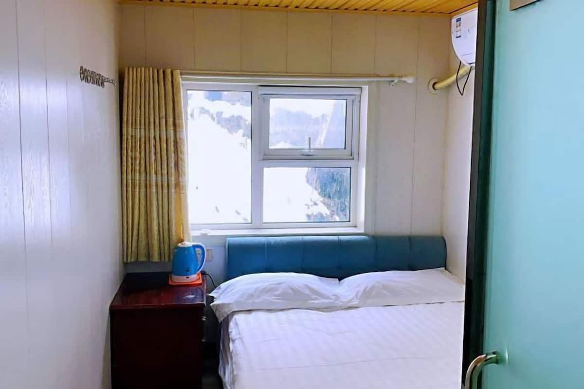 Una habitación con una cama, una ventana con cortinas, una mesita de noche con hervidor y un calentador fijado a la pared.