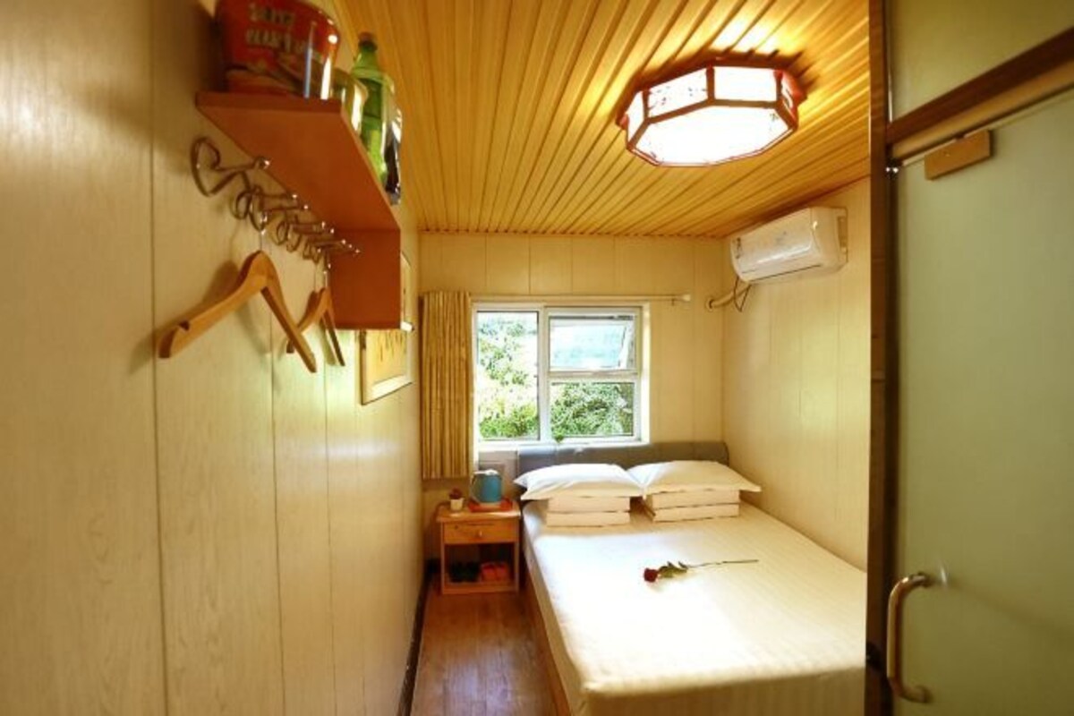 A compact room with a bed, a window, a bedside table, and a shelf with items.