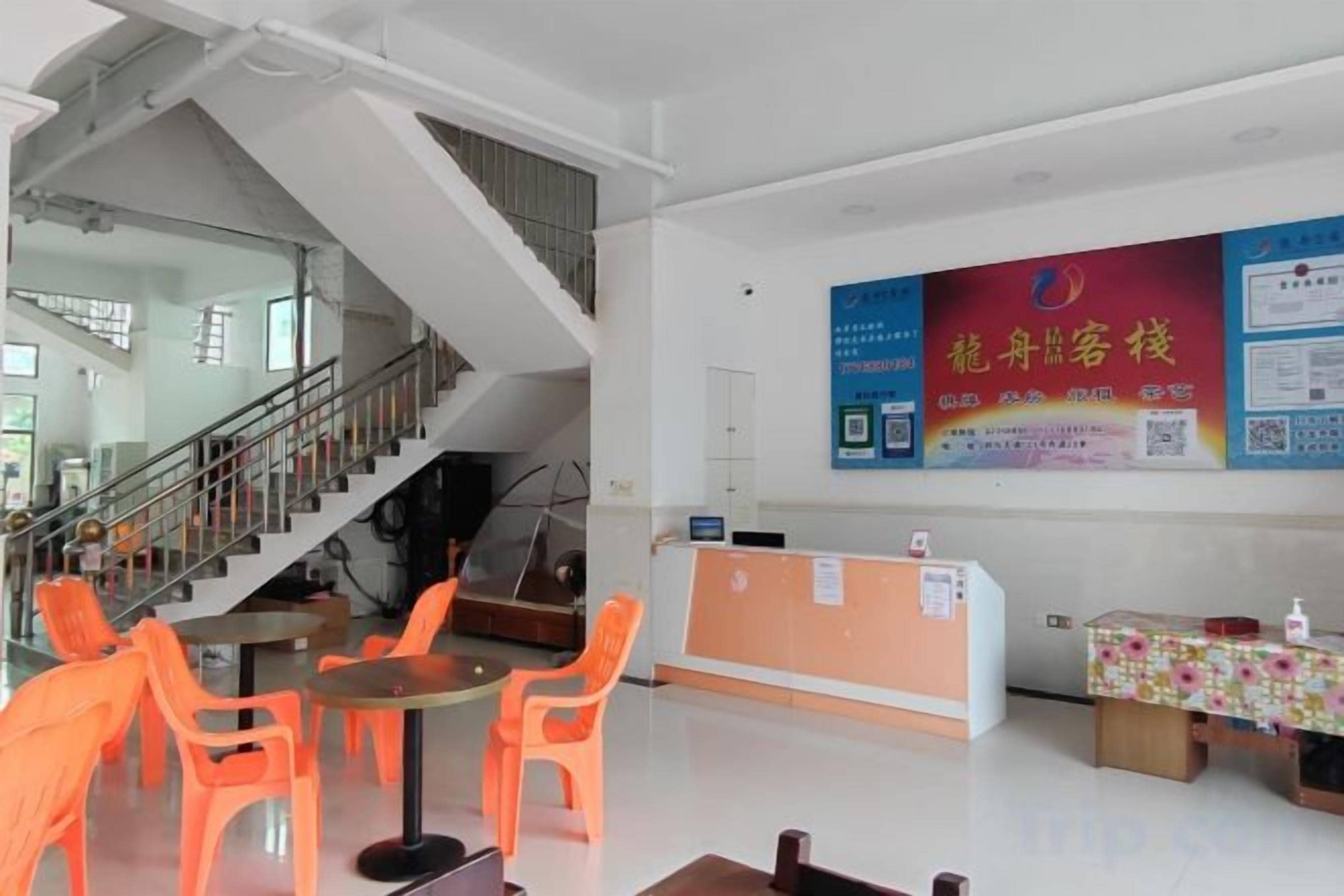 A hotel lobby with a reception desk, orange chairs, a staircase, and a large poster on the wall.