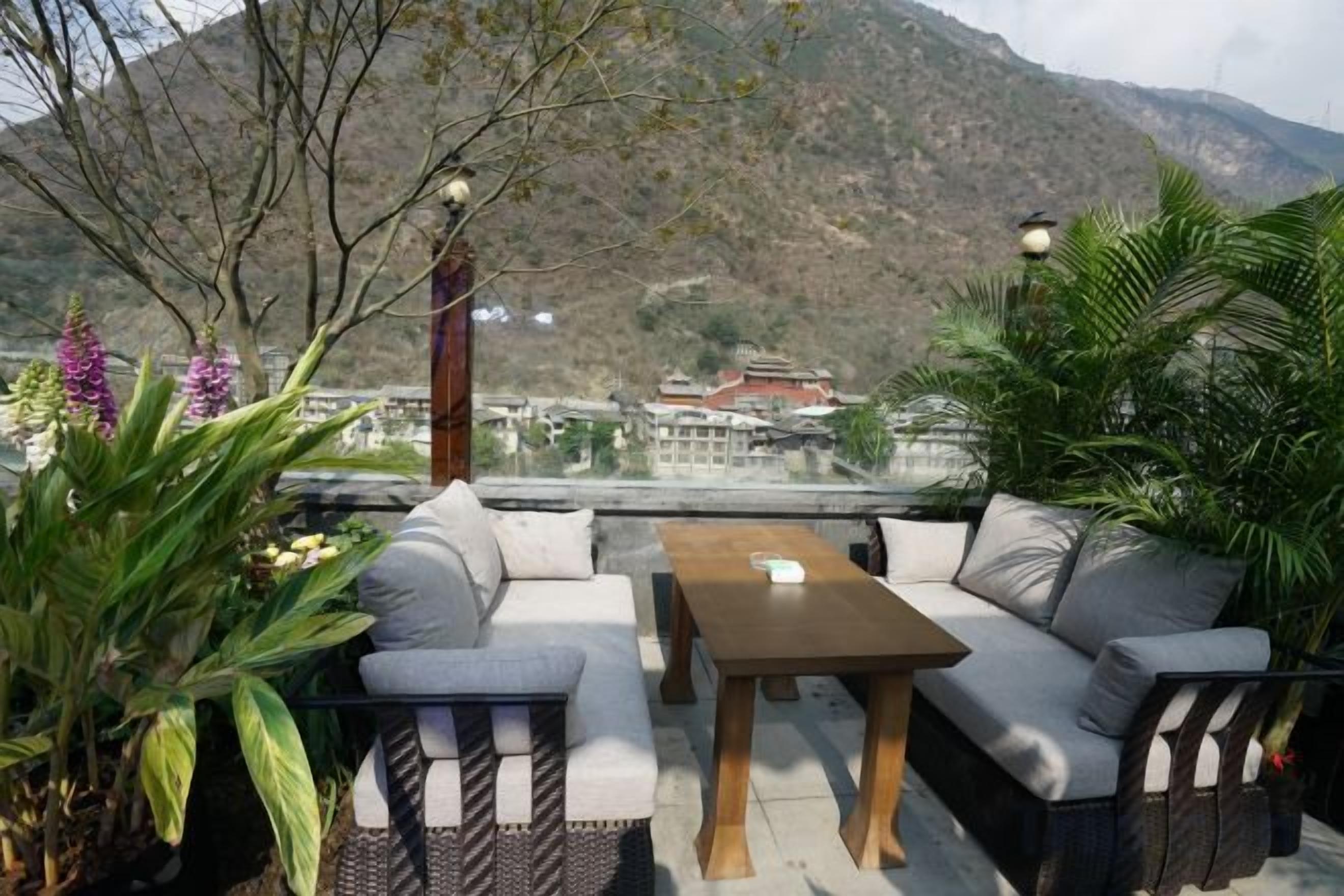 An outdoor seating area with a sofa and table, surrounded by plants, overlooking a mountainous landscape.