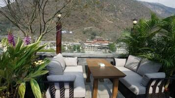 An outdoor seating area with a sofa and table, surrounded by plants, overlooking a mountainous landscape.