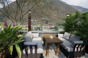 Área externa com assentos, incluindo um sofá e mesa, cercada por plantas, com vista para uma paisagem montanhosa.