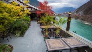 Una terraza al aire libre con mesas y sillas, rodeada de abundante vegetación y vistas a un río y montañas.