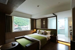 Una habitación de hotel moderna con una cama grande, un pequeño rincón de descanso, una mesita de noche, una lámpara y vistas al exterior con zonas verdes.