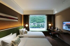 Habitación de hotel con dos camas, un televisor, un escritorio y una ventana con vistas a zonas verdes y edificios.