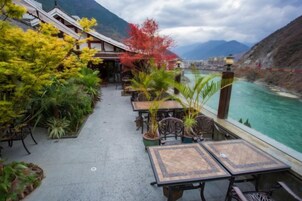 Una terraza con mesas y sillas que da a una extensión de agua, rodeada de vegetación y con montañas al fondo.