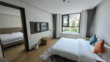A modern hotel room with a large bed, a TV mounted on the wall, a window with a view of greenery, and a small wooden side table.