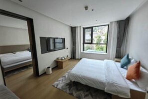 Una habitación de hotel moderna con una cama grande, un televisor montado en la pared, un ventanal con vistas a zonas verdes y una mesita de noche de madera.