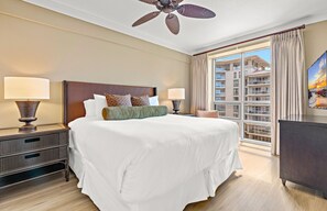A hotel room with a large bed, two bedside lamps, a ceiling fan, a window with curtains, and a view of a building.