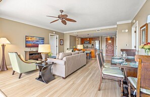 A spacious living room with a sofa, chairs, a dining table with chairs, a kitchen area, and a ceiling fan.