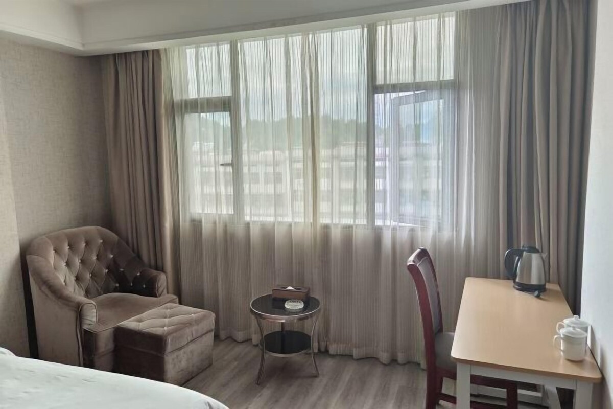 A hotel room with a bed, a tufted armchair, a small round table with a kettle, a chair, and a view through sheer curtains.