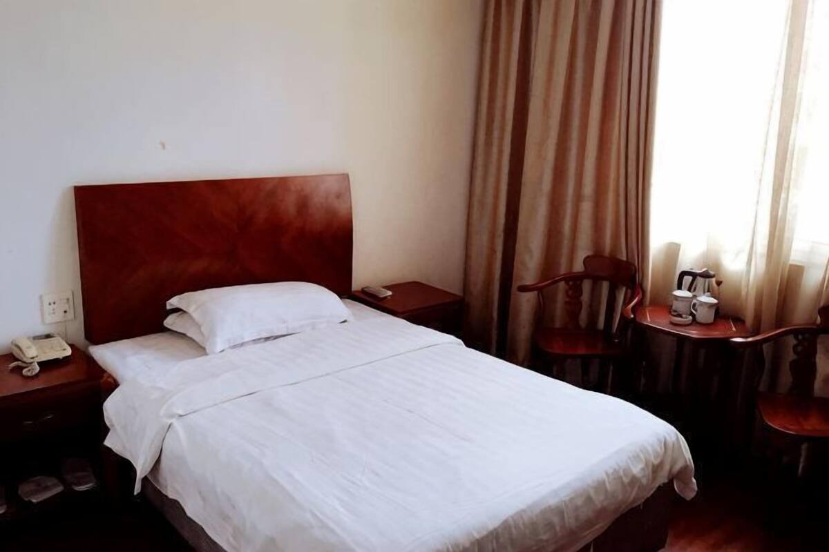 A hotel room with a wooden headboard bed, a bedside table with a phone, a chair, and a small table with a cup and saucer.