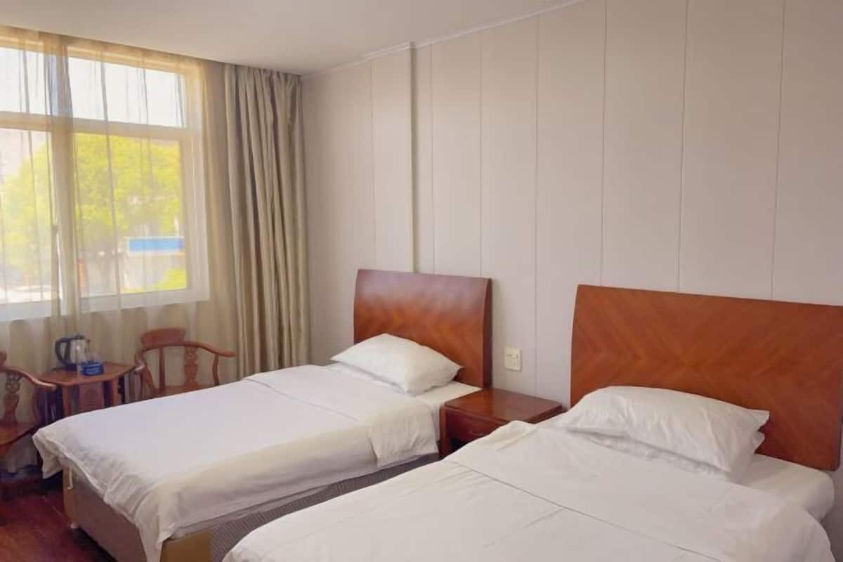 A hotel room with two beds, a wooden headboard, a small table with a kettle, and a window with curtains.