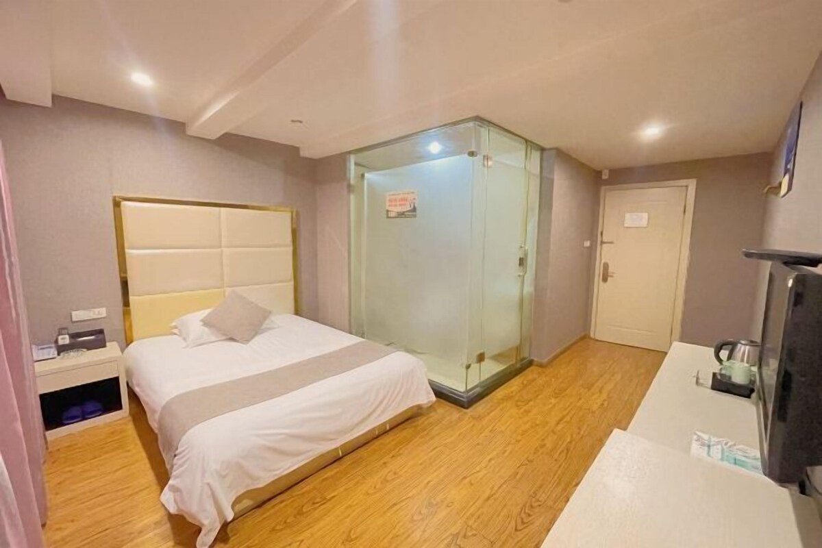 A hotel room with a bed, a glass-enclosed shower, a white door, and a small table with a kettle.