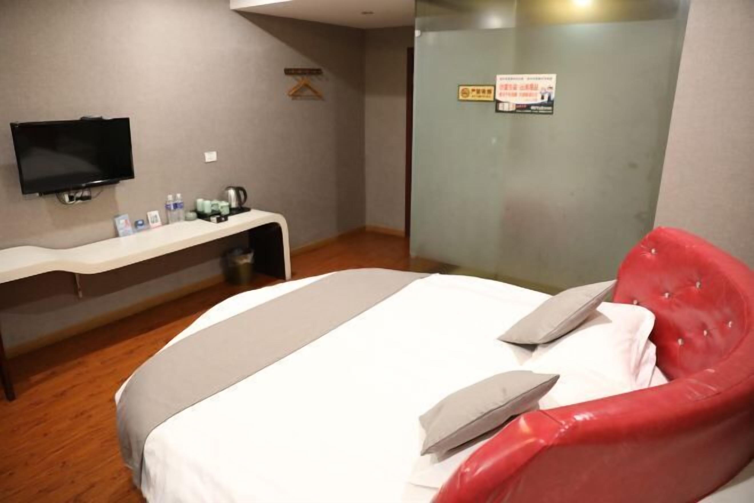 A hotel room with a bed, a TV mounted on the wall, a desk with toiletries, and a red upholstered chair.