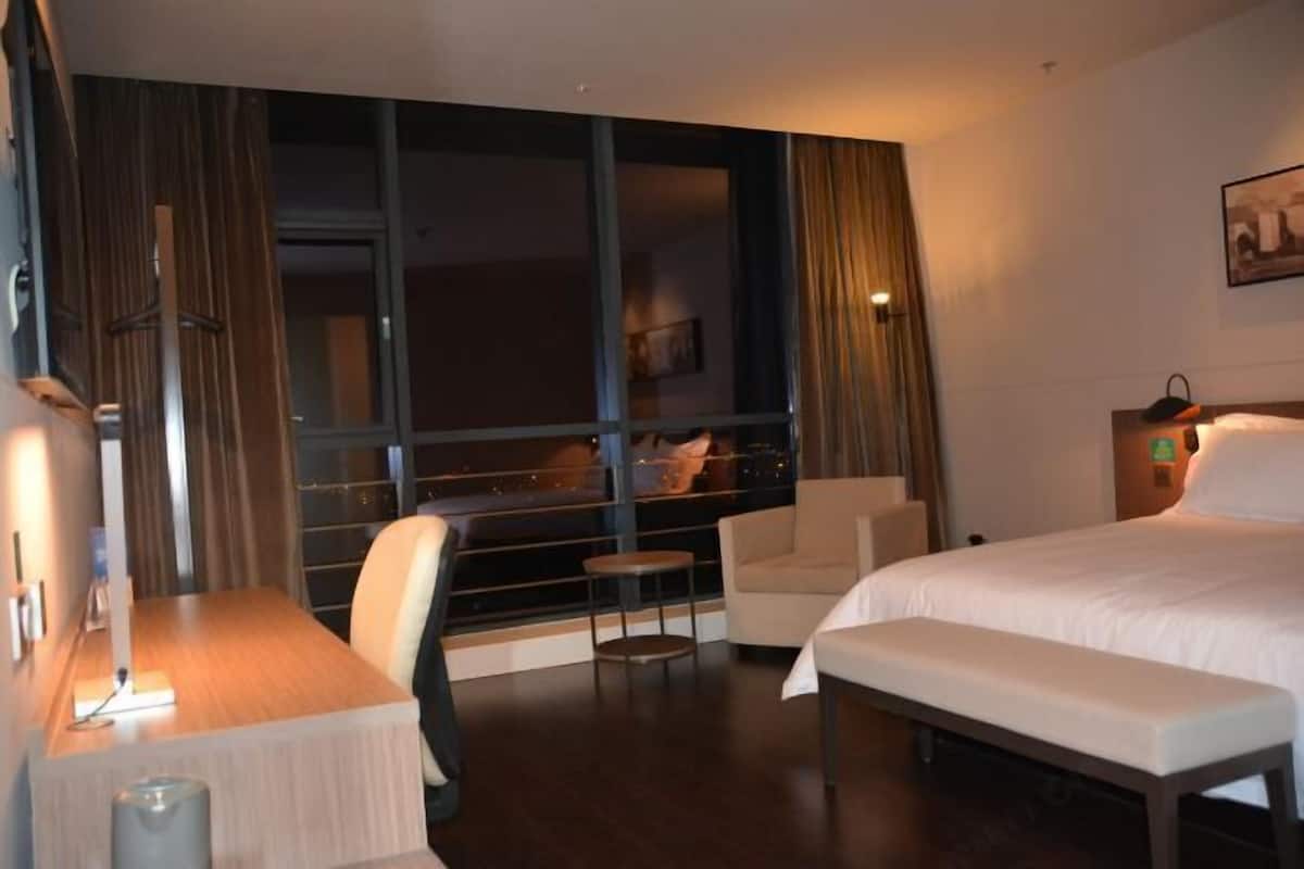 Une chambre d’hôtel équipée d’un lit, d’un bureau, d’une chaise, d’une petite table et offrant une vue sur le paysage urbain grâce à de grandes fenêtres.