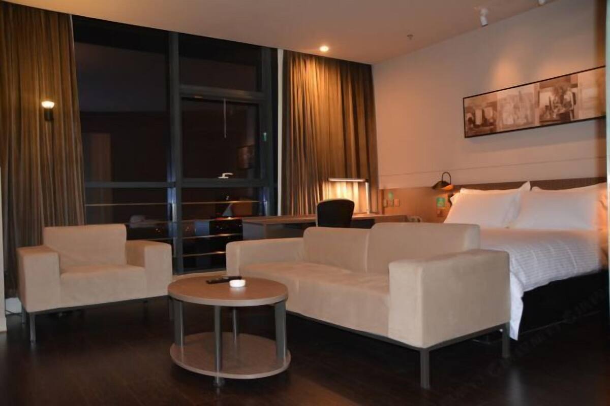 Une chambre d’hôtel avec un lit, deux canapés blancs, une petite table ronde, un bureau avec une lampe et une grande fenêtre avec des rideaux.