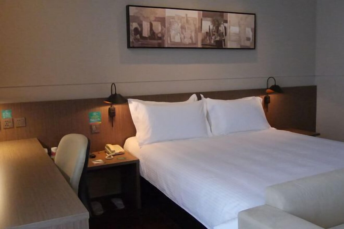 A hotel room with a bed, bedside table, lamp, chair, and a painting on the wall.