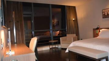 A hotel room with a large window, a bed, a desk, a chair, and a bench.