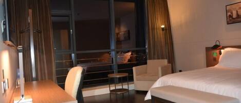 A hotel room with a large window, a bed, a desk, a chair, and a bench.