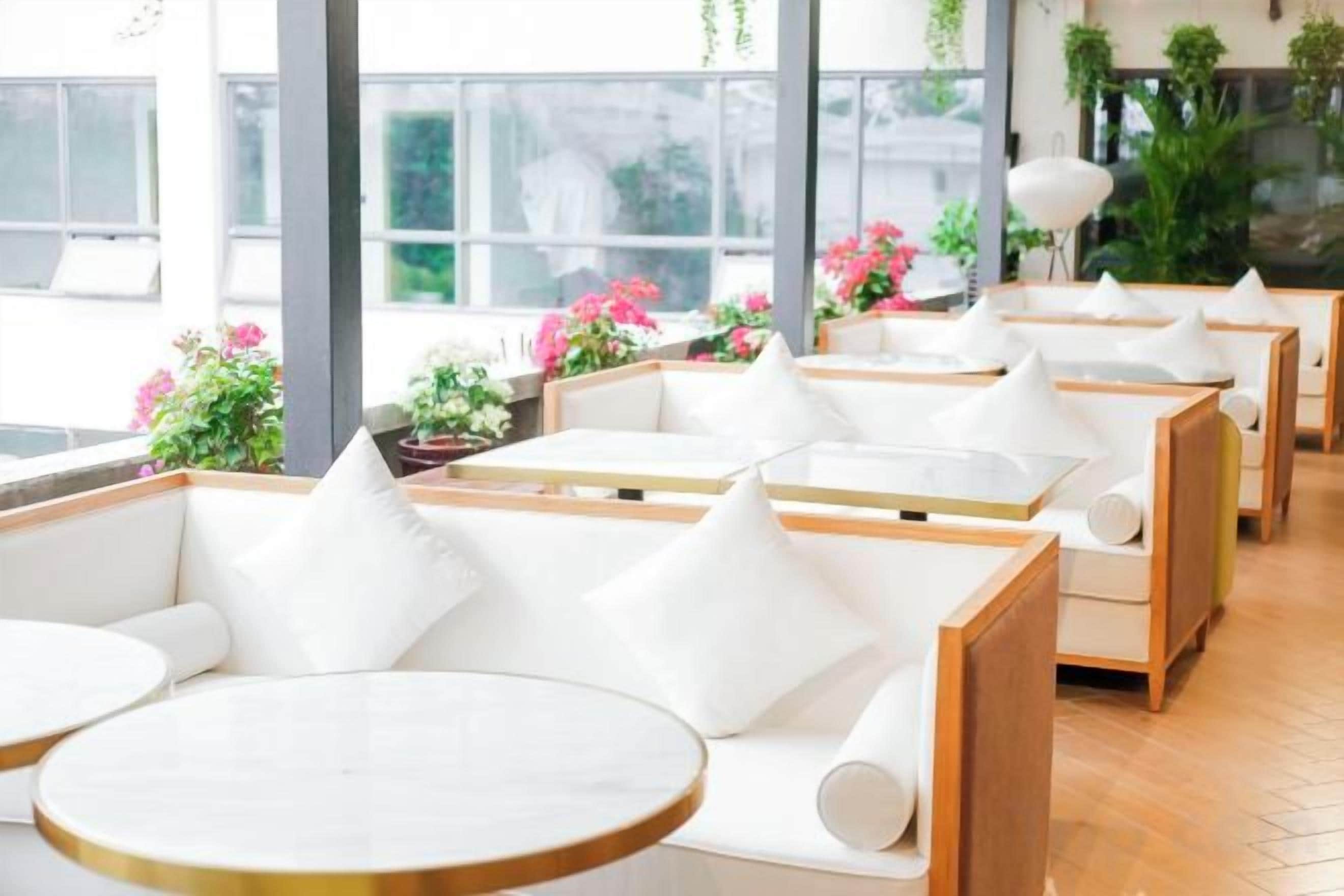 A modern interior with white cushioned seating and round tables, large windows, and indoor plants.
