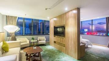 Quarto de hotel moderno com uma televisão grande presa a um painel de madeira, uma área de estar acolhedora com um sofá e poltronas, uma pequena mesa lateral e vista para o horizonte noturno.