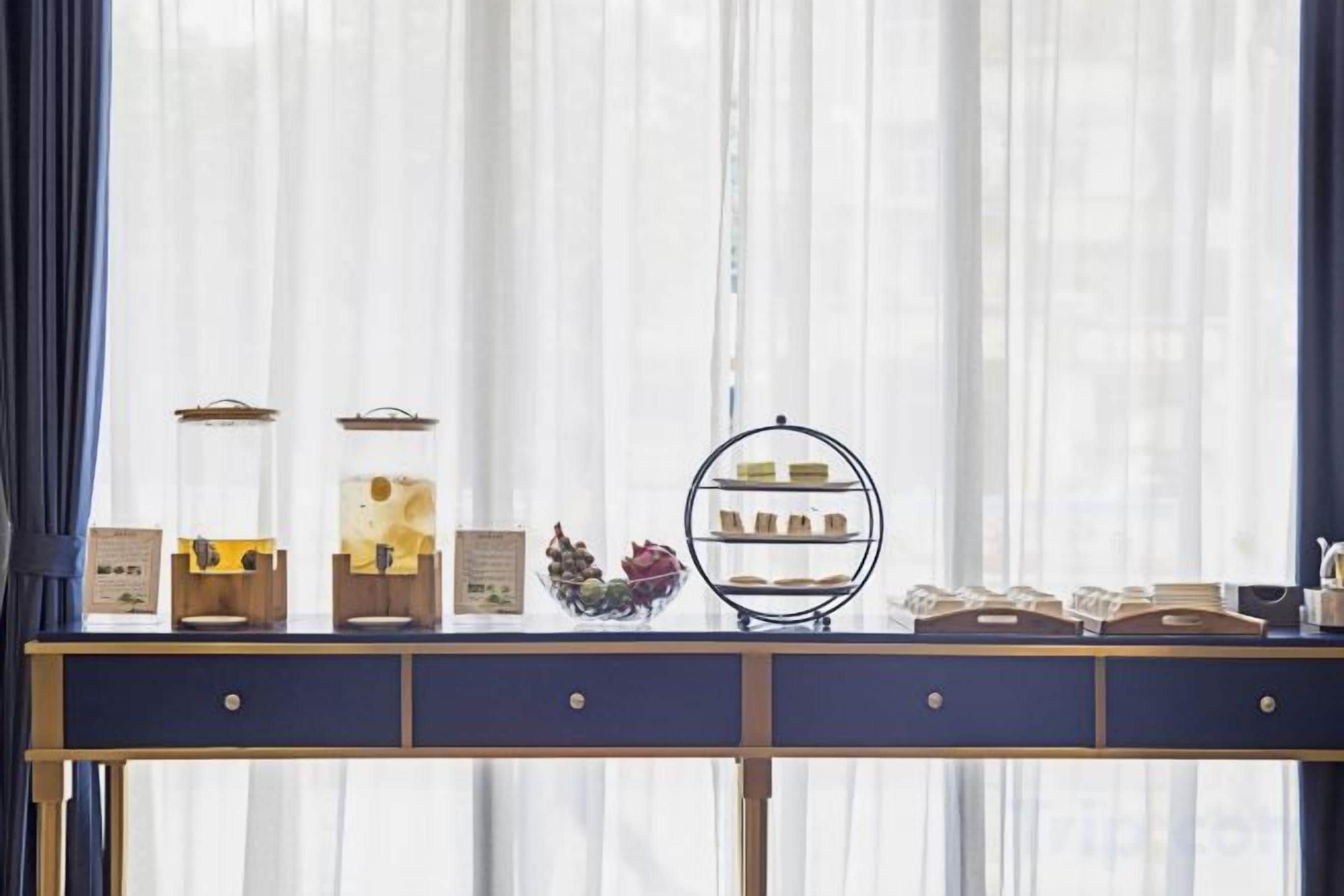 A table with various food items and a display stand in front of a window with sheer curtains.