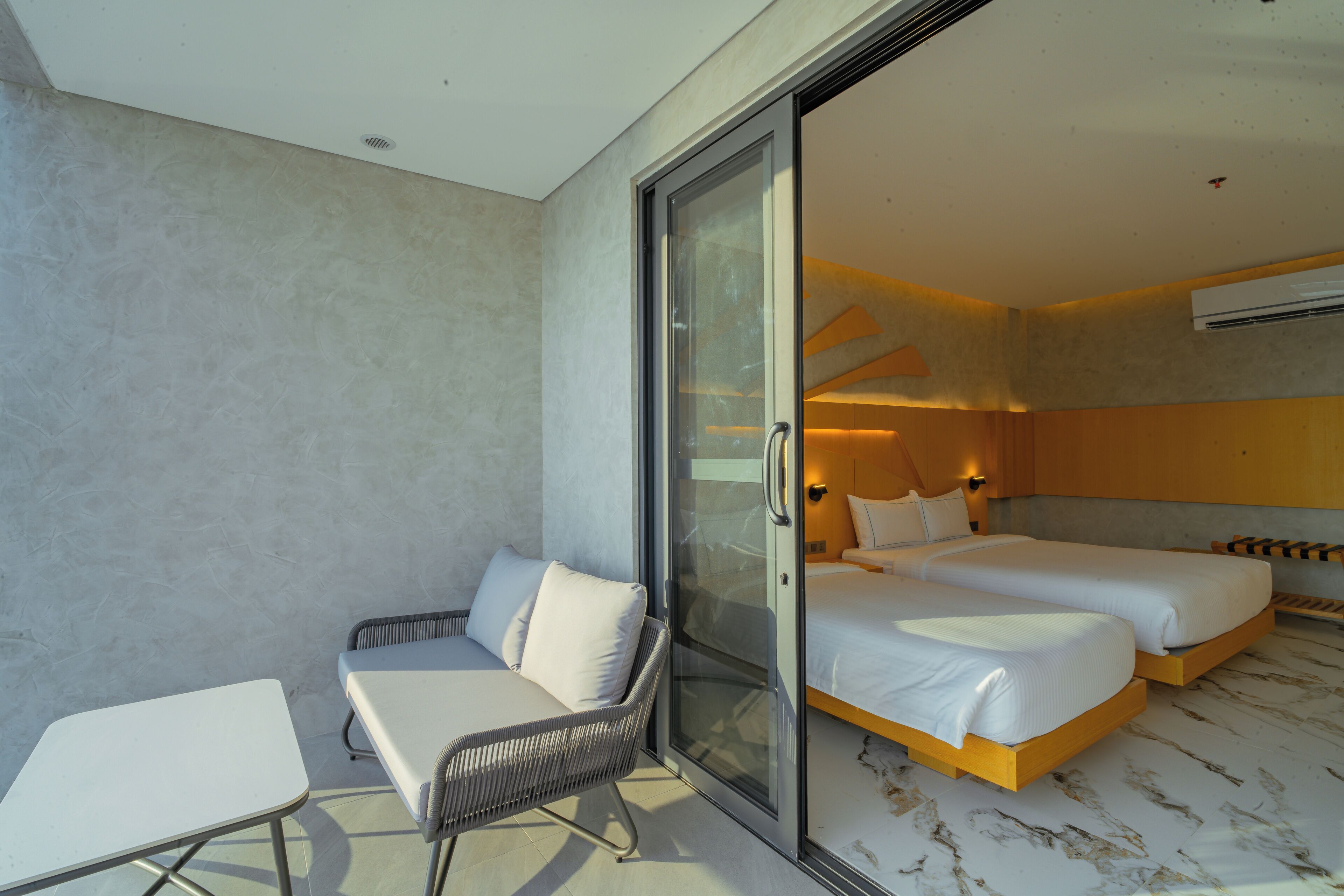 Quarto de hotel moderno com porta de vidro que dá para uma varanda, um sofá, duas camas e uma mesa pequena.