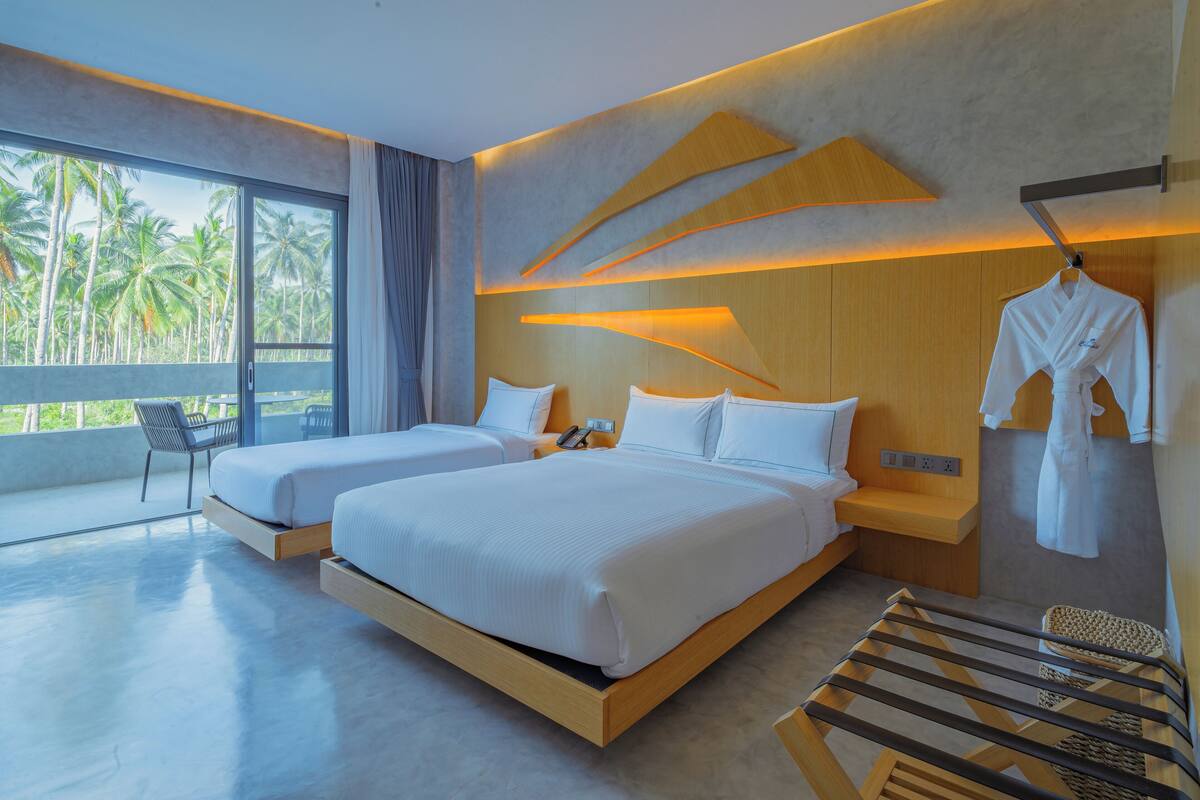 Une chambre d’hôtel moderne avec deux lits, un balcon donnant sur des palmiers et une robe accrochée au mur.