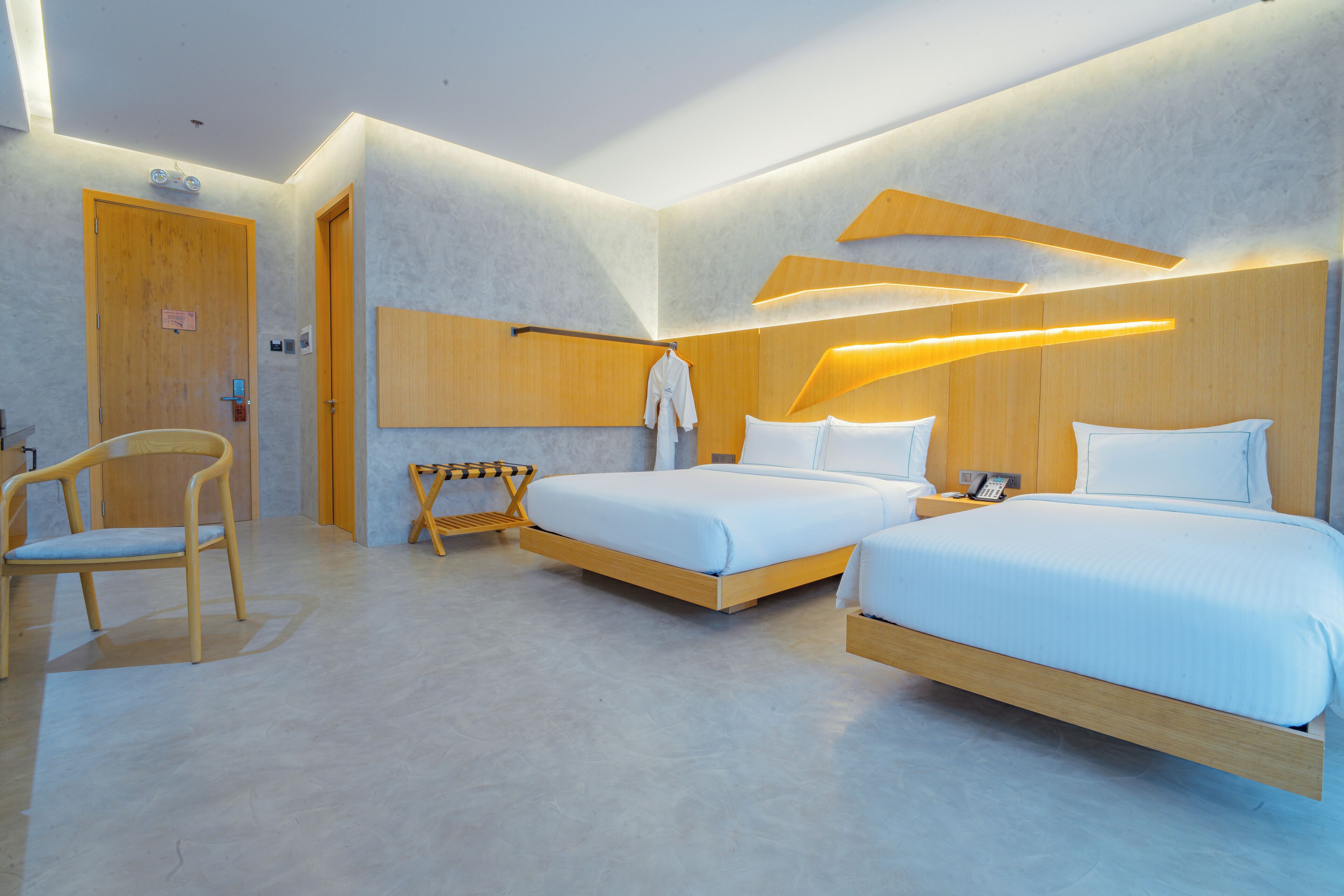 A modern hotel room with two beds, a wooden chair, a small table, and a robe hanging on the wall.