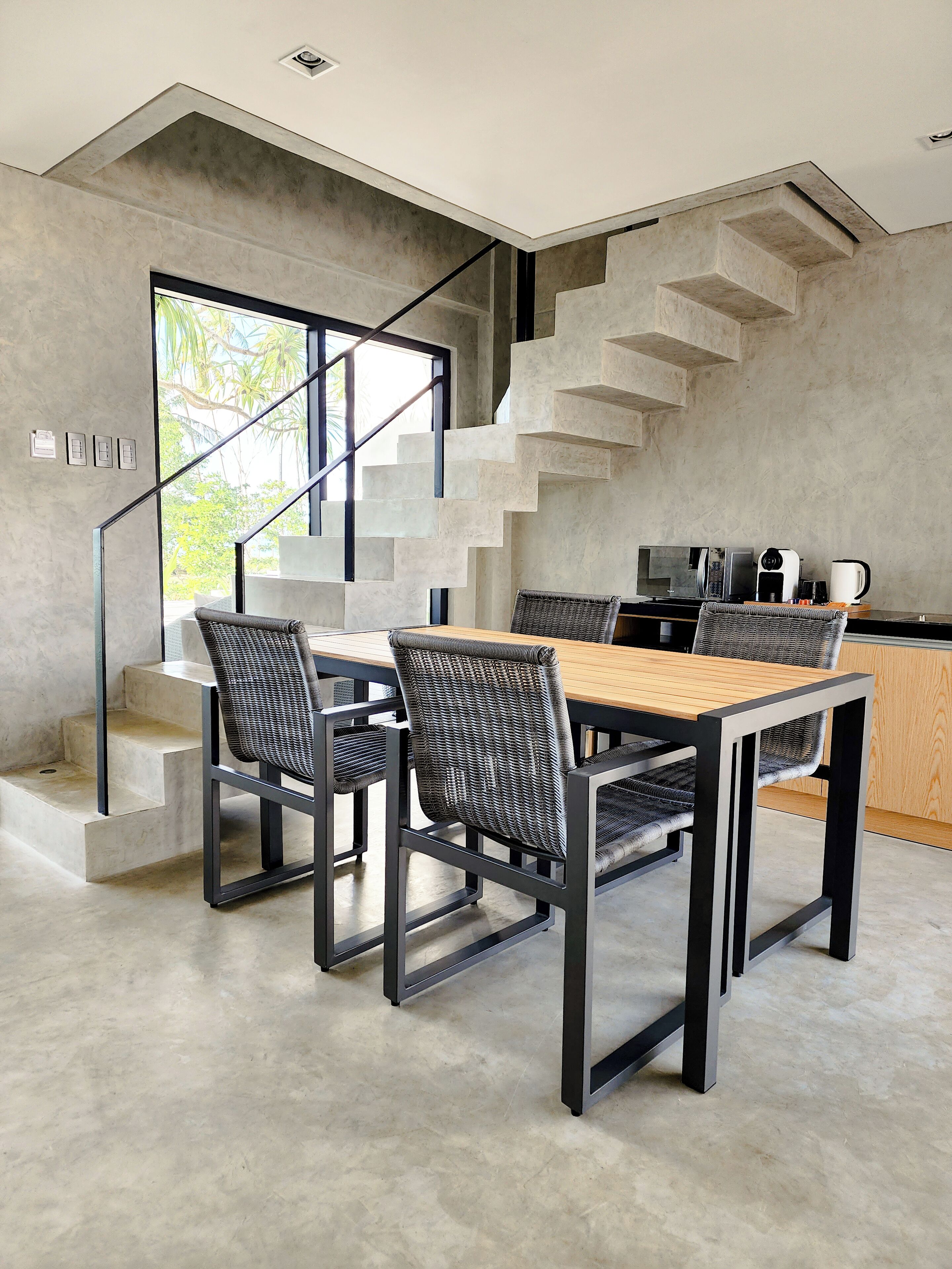 A modern kitchen with a wooden dining table and chairs, a staircase, and a view of greenery outside.