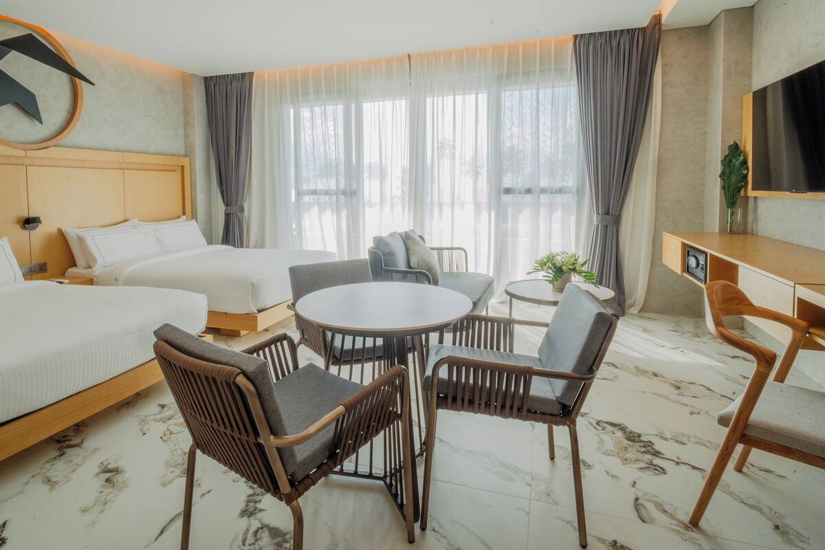 Une chambre d’hôtel avec deux lits, une table à manger avec des chaises, une télévision et une grande fenêtre avec des rideaux.