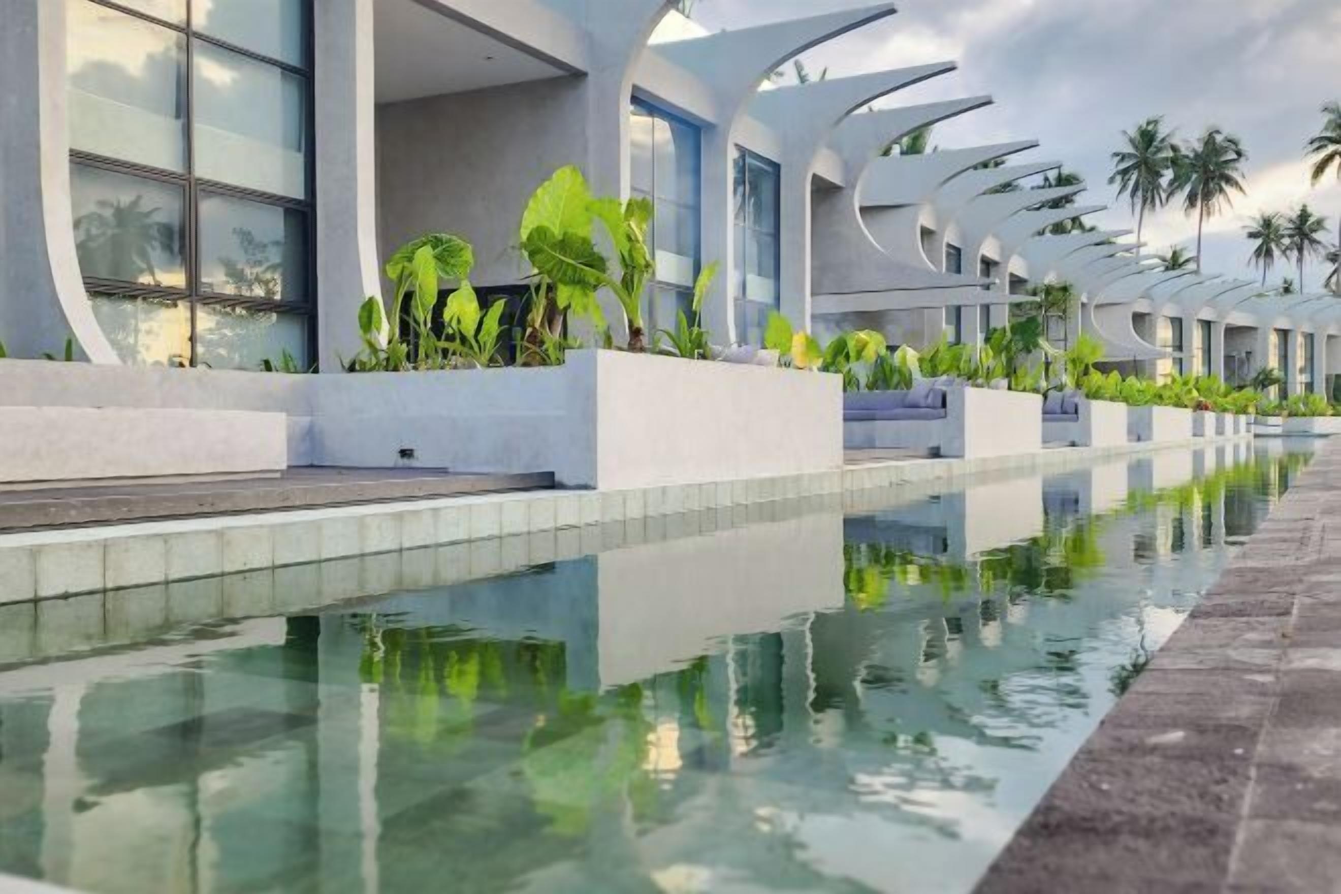 A modern hotel with a long pool, outdoor seating, and tropical plants.
