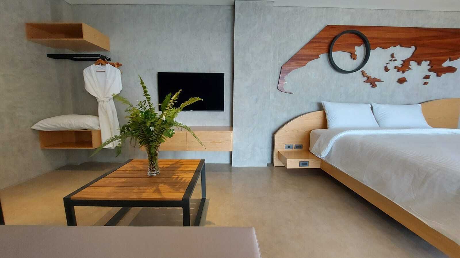 A modern bedroom with a wooden bed, a small table with a plant, a TV, and a wall-mounted shelf with a robe.