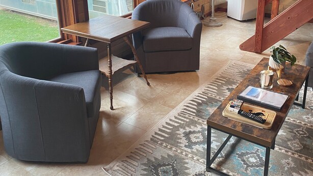 A cozy living room with a large window, a wooden table, a grey armchair, and a coffee table with books and a remote control. - Red Bluffs Lodge Upper Unit, located on the River, minutes from Devils Tower! (Hulett)
