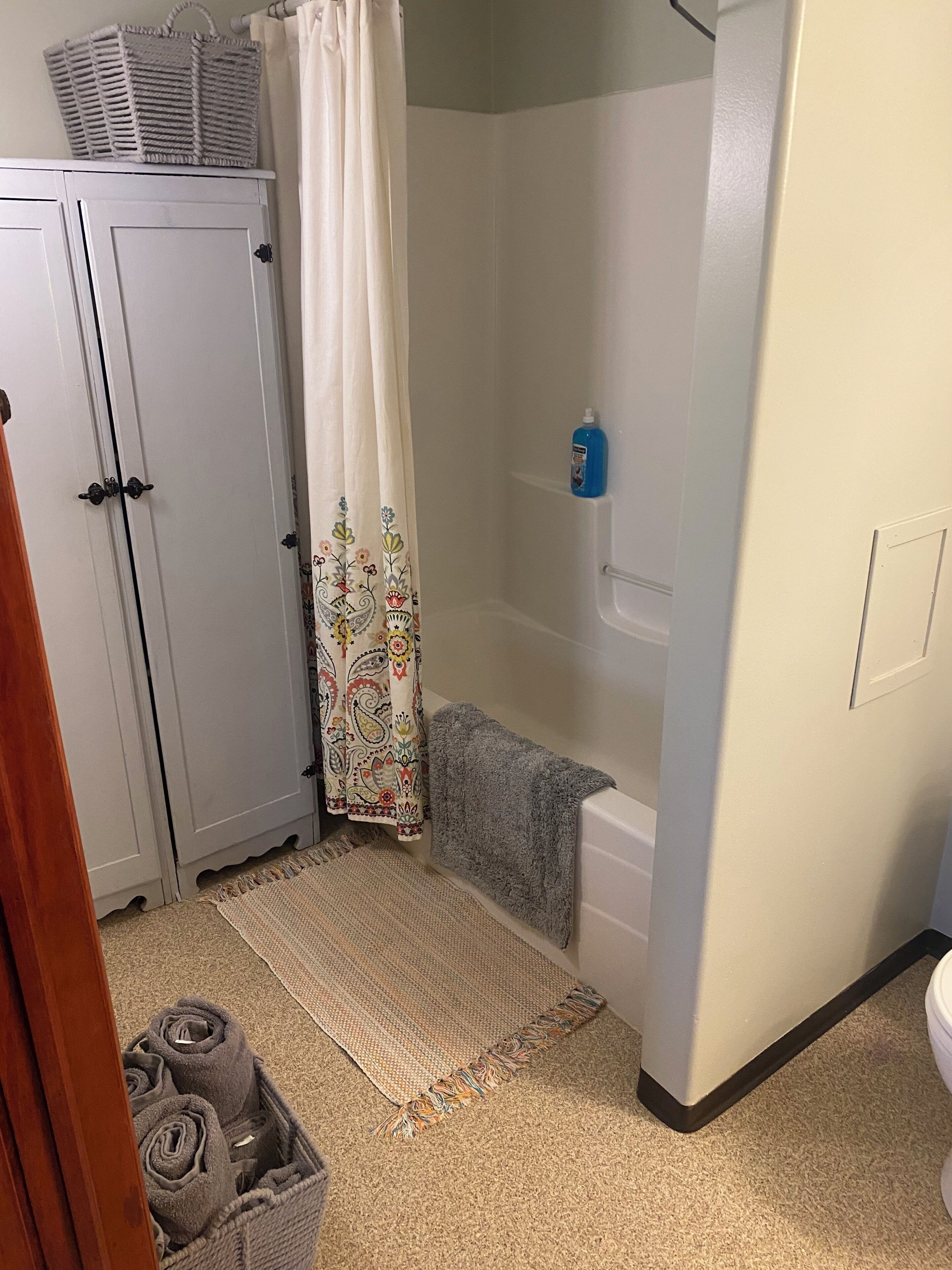 A bathroom with a shower, a toilet, a sink, a white cabinet, a patterned shower curtain, and a rug.