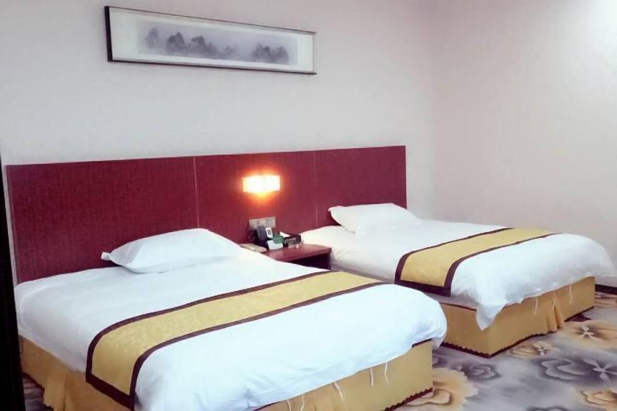 A hotel room with two beds, each with a white bedspread and a yellow and brown striped blanket. There is a framed picture on the wall above the beds and a nightstand with a lamp between them.