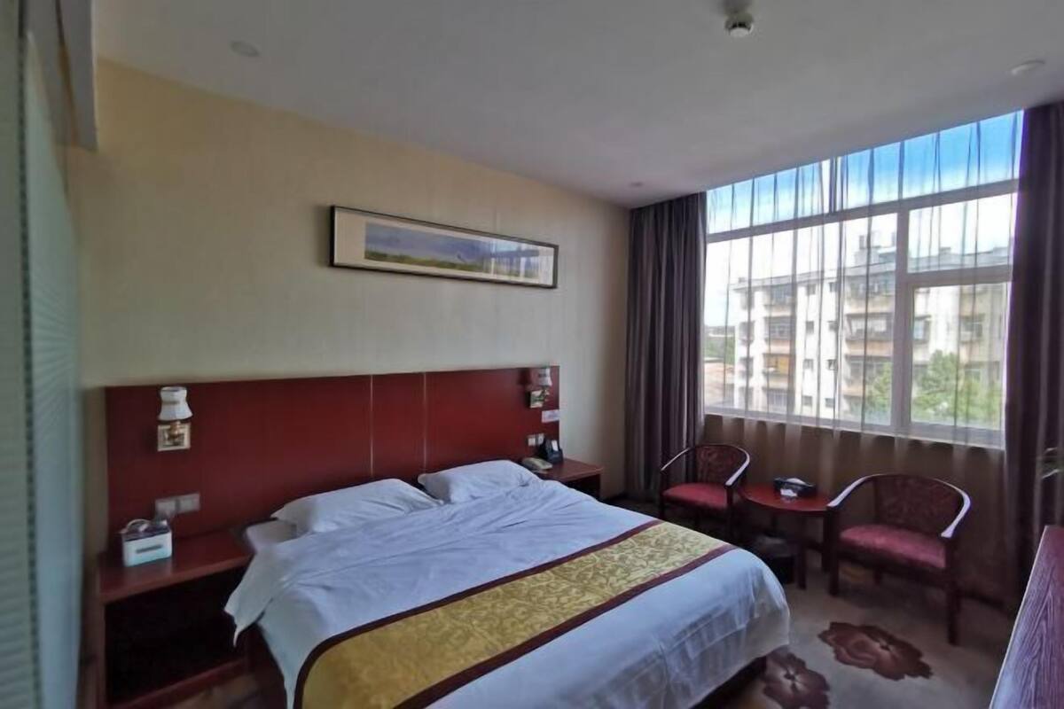 A hotel room with a large bed, a desk, two chairs, a window with a view of buildings, and a painting on the wall.